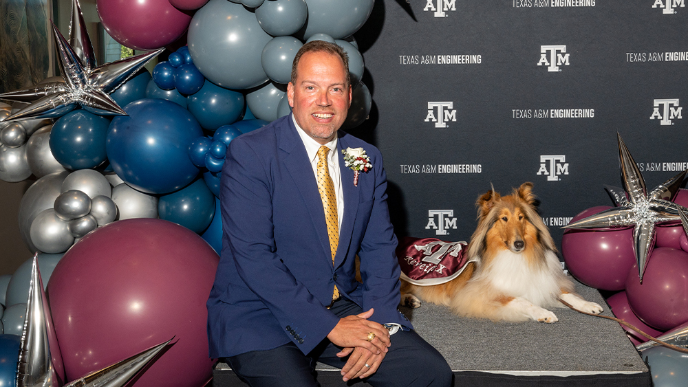 A man in a suit sits with the Texas A&M mascot, Reveille, in front of a maroon backdrop and balloons