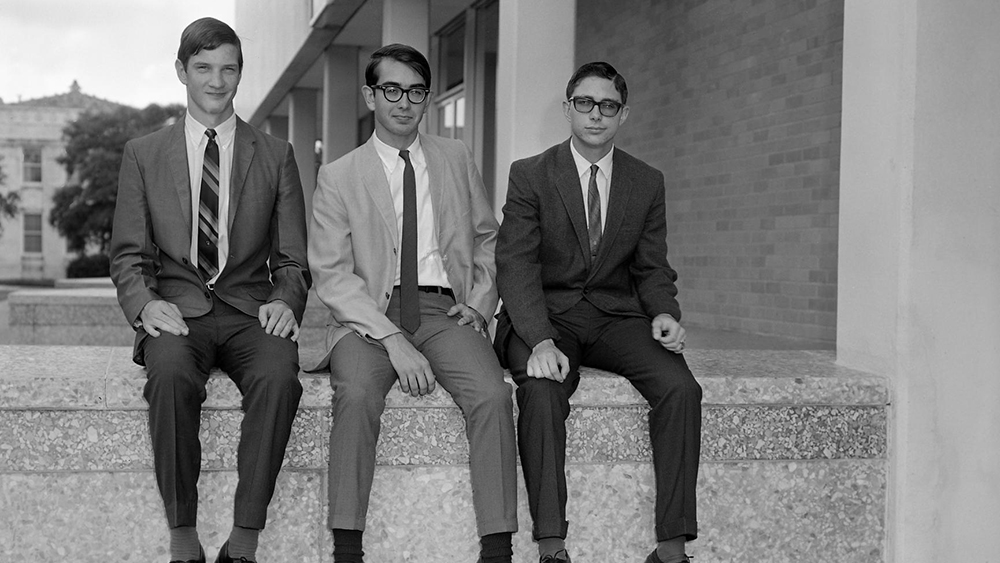 Black and white photo of three individuals in suits sitting on a concrete wall.