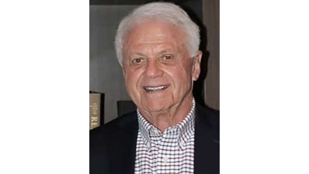 A smiling man with white hair wearing a dark jacket and checked shirt