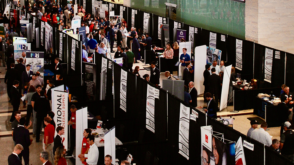 A large group of individuals move among booths within a large event space.
