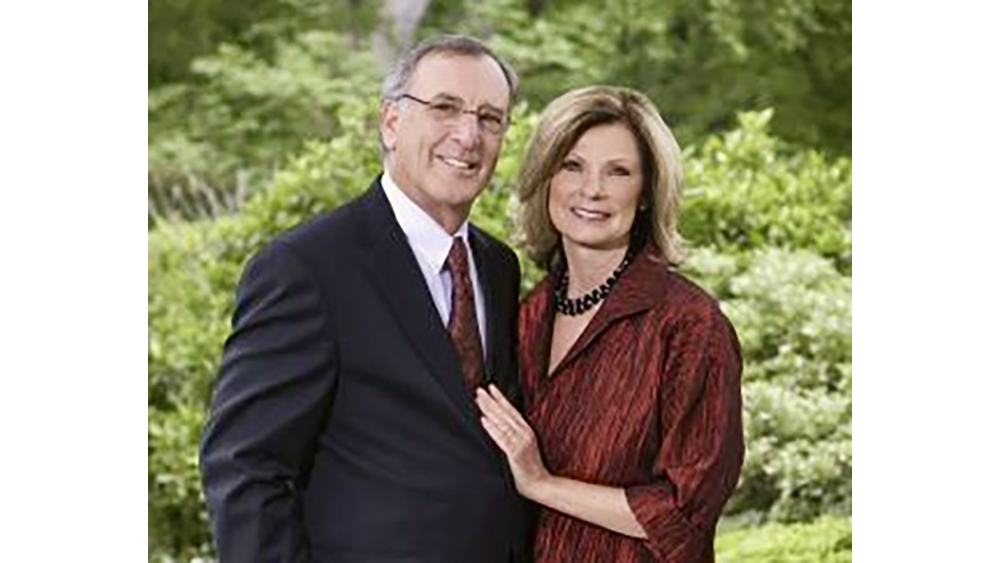 A man and a woman in business attire pose in front of green bushes.