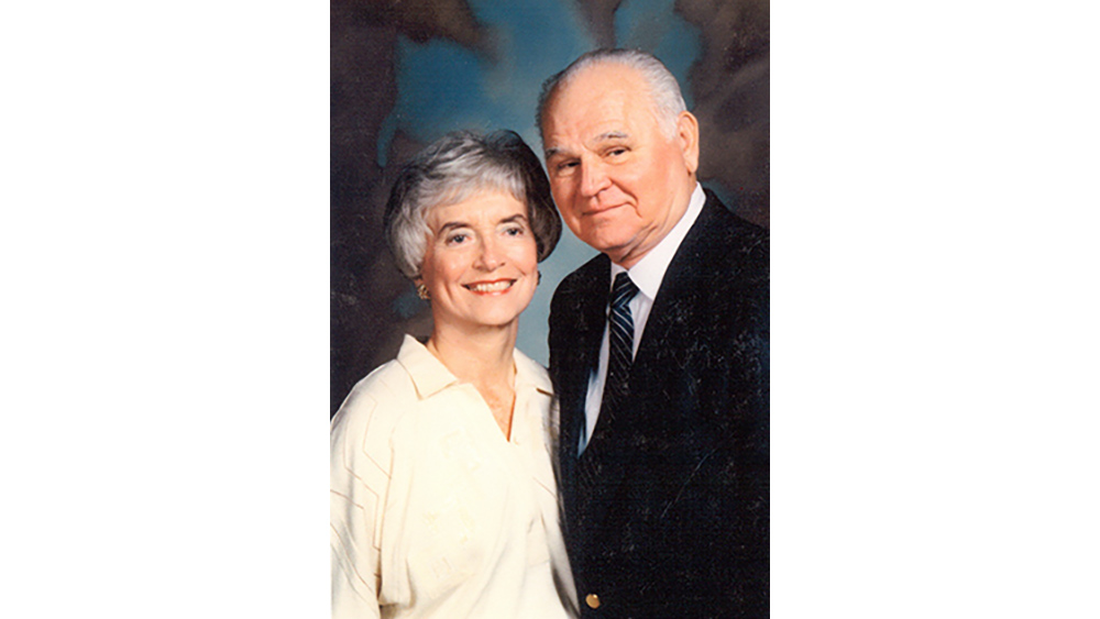 A woman in a cream blouse and a man in a dark suit pose together in front of a blue background.