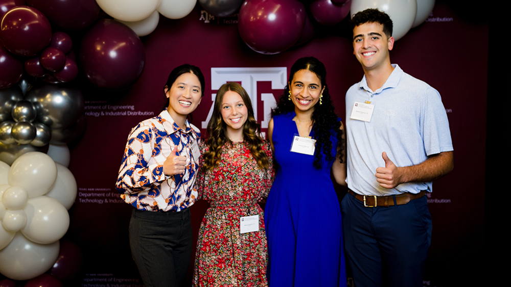 Four individuals in business attire smile while standing in front of a maroon backdrop