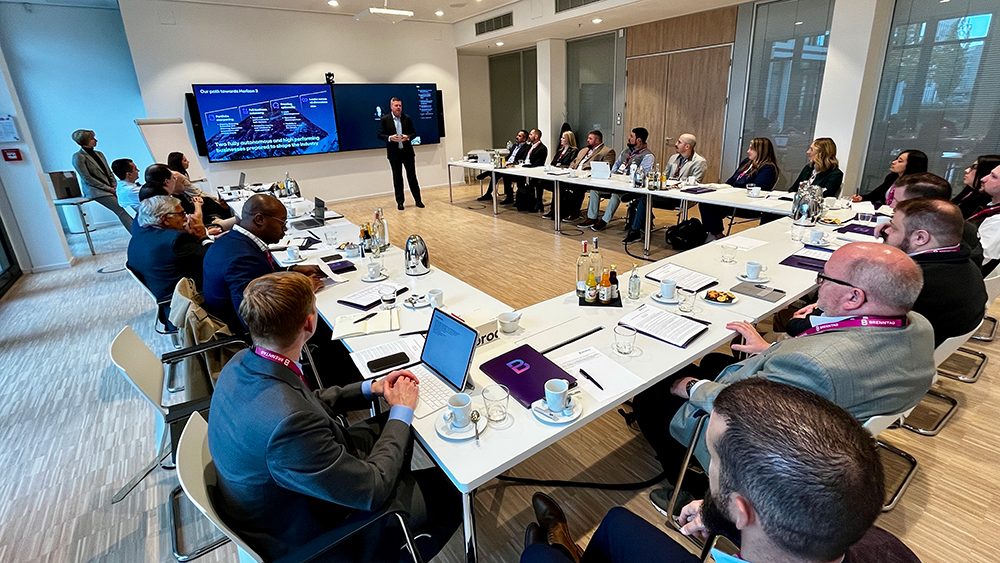 A wide view of a large room where over 20 people seated around a U-shaped table are listening to a standing person speak.