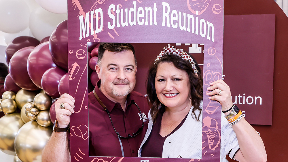 A man and a woman smile and hold up a decorated cardboard frame with the words, "MID Student Reunion" on it.