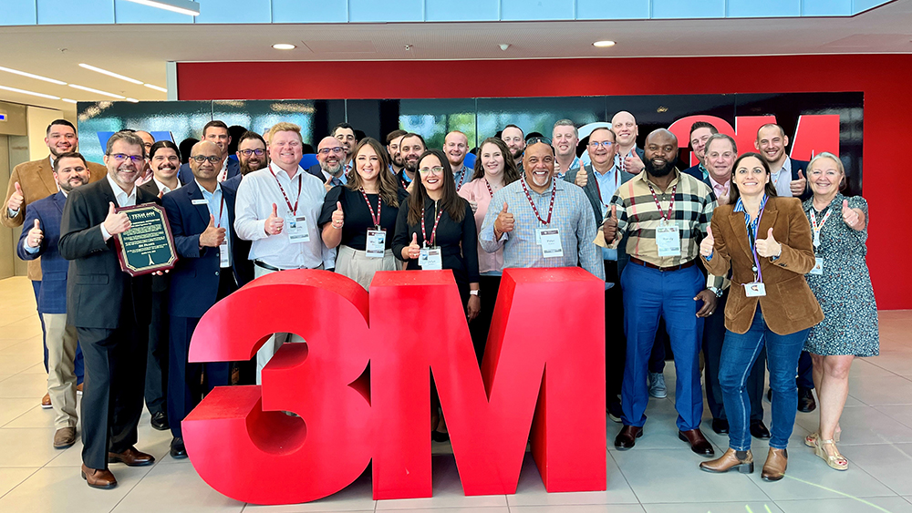 A crowd of smiling people standing behind a waist-high three-dimensional 3M company logo in the middle of a large lobby
