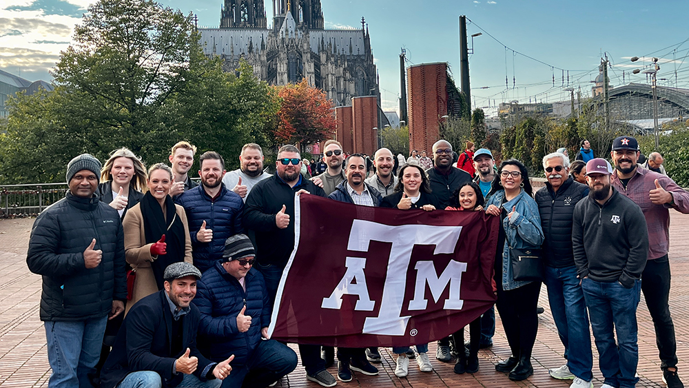 Group of smiling people standing together in front of a large stone church while holding a large flag with the Texas A&amp;M University text logo (ATM) on it.
