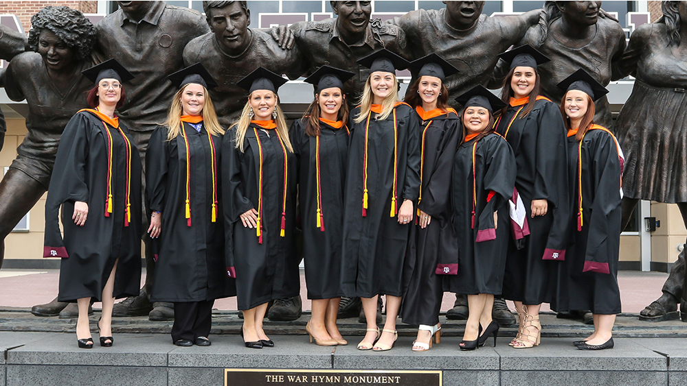 Nine people wearing graduate-degree-level graduation robes and mortarboards smile as they stand on the base of a large statue labeled, "The War Hymn Monument."