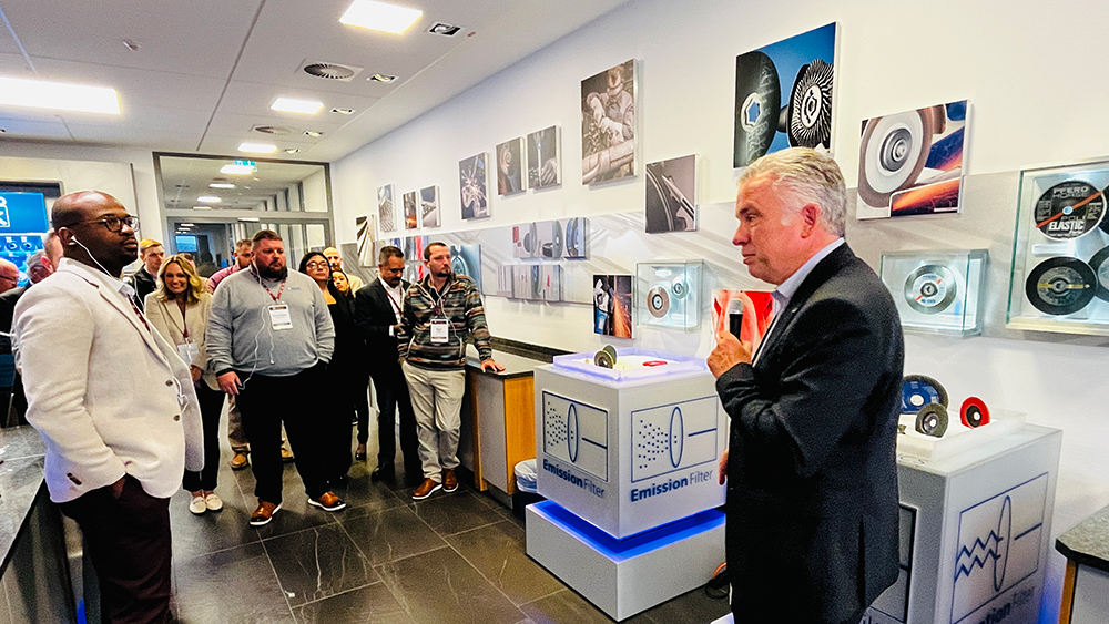 A man in business attire speaks to visitors in a lobby area decorated with images and displays of company products. One kiosk is labeled, 'Emissions Filter."