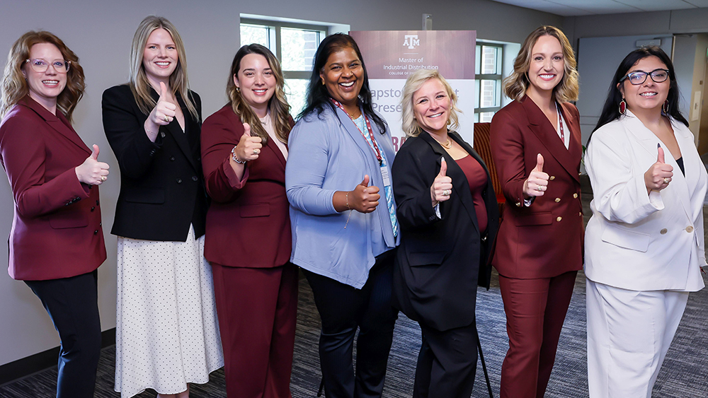 Seven smiling women in business attire standing in front of a tall banner featuring the Master of Industrial Distribution Program at Texas A&amp;M University logo. 