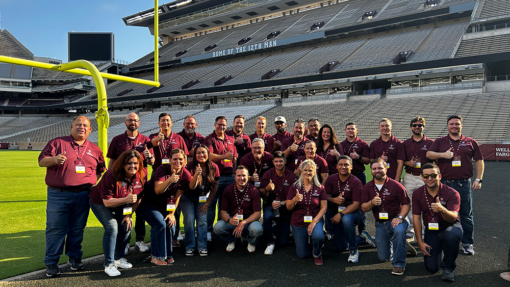 A large group of smiling people posing for a picture near a goalpost in the Texas A&amp;M University football stadium. Words behind them read: Home of the 12th Man.