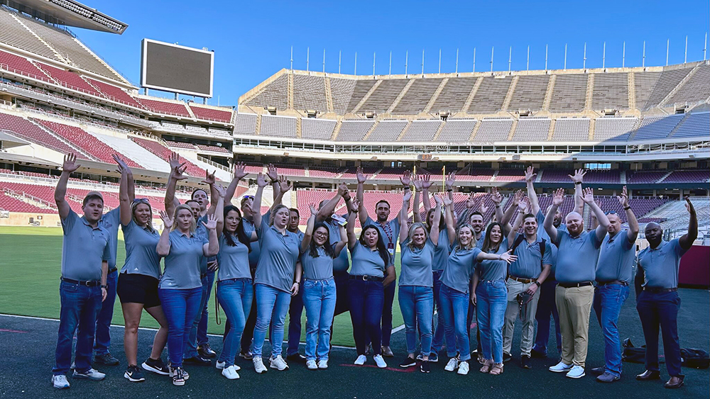 A large group of people with their hands and arms raised cheer while standing on the field of a huge, empty football stadium.