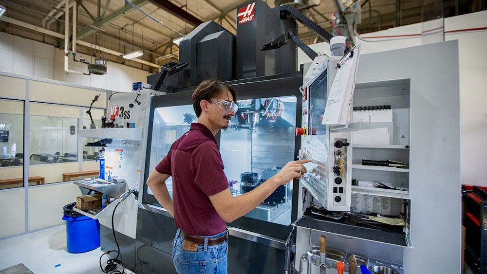 A student wearing safety glasses pushes a button on a large machine in a lab.