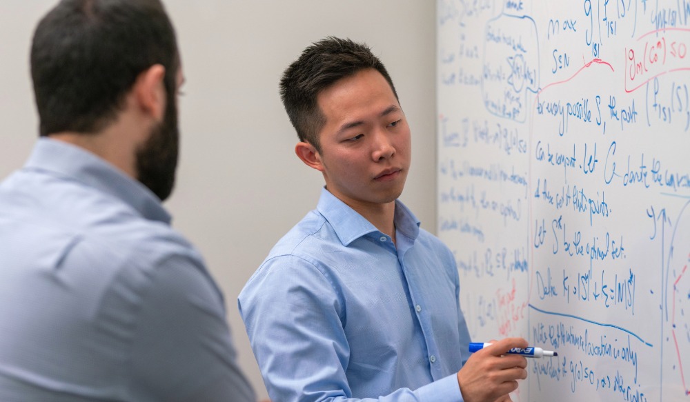 A researcher writes out an optimization proof on a whiteboard while a colleague looks on
