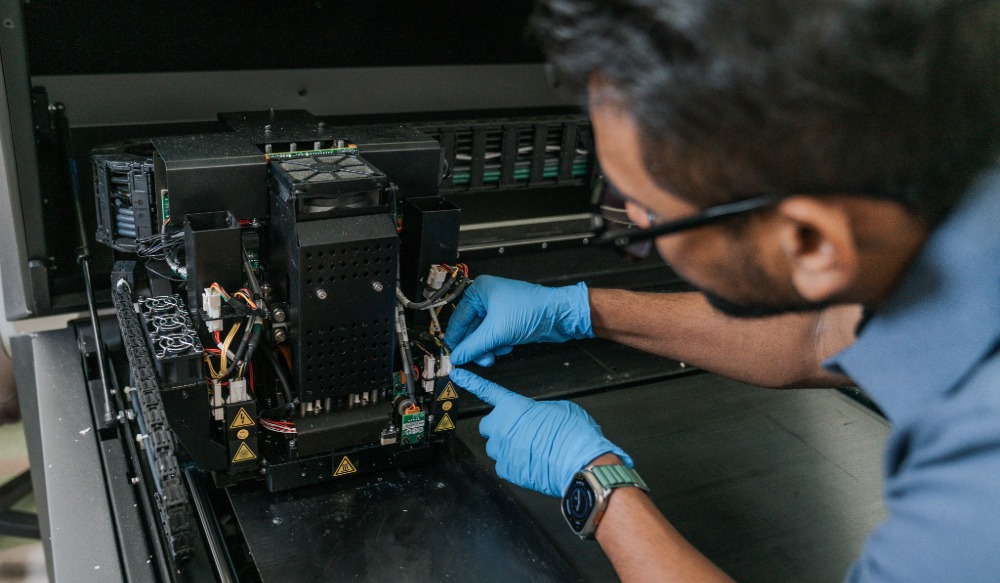 A researcher wearing gloves works on additive manufacturing equipment in a lab, adjusting internal components.