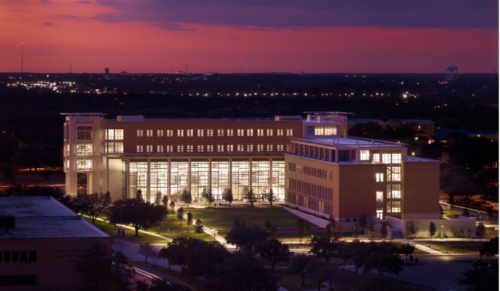 Aerial twilight view of the Engineering Technology Building (ETB) on the Texas A&amp;M University campus, illuminated from within, with landscaped grounds and a pink-purple sunset sky