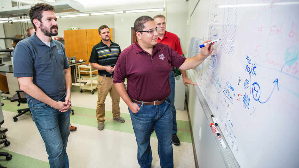 Professor drawing diagrams and writing mathematics on the white board with three students standing behind him