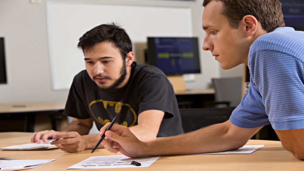 Two ISEN students in a classroom lean over a desk, pointing at a printed diagram while discussing coursework together.