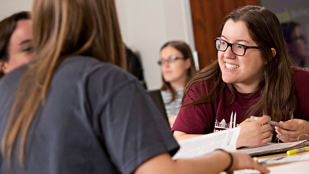 A smiling ISEN student in a maroon T-shirt discusses coursework with classmates in a busy classroom setting