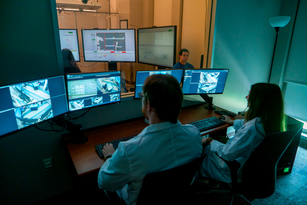 Two people in lab coats sit in a control room in front of multiple computers showing camera angles. They are facing a window, where there is another room with two people conducting experiments surrounded by large screens of information. 