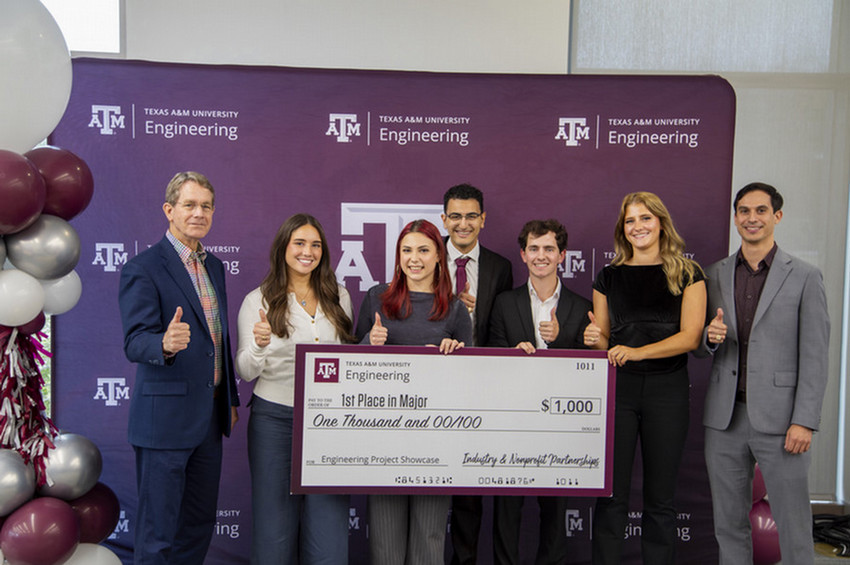 A group of seven individuals poses in front of a Texas A&amp;M University Engineering backdrop, holding a large check. The check is labeled "1st Place in Major" with an amount of $1,000. The group consists of five students and two adults, all smiling and giving thumbs-up gestures.