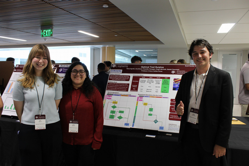 Three students stand in front of a research poster at an academic conference. The poster features diagrams and data related to their project, titled "Optimal Tool Surplus." The setting is a well-lit indoor space with other attendees visible in the background. The students are smiling, dressed in business-casual attire, and wearing name tags.