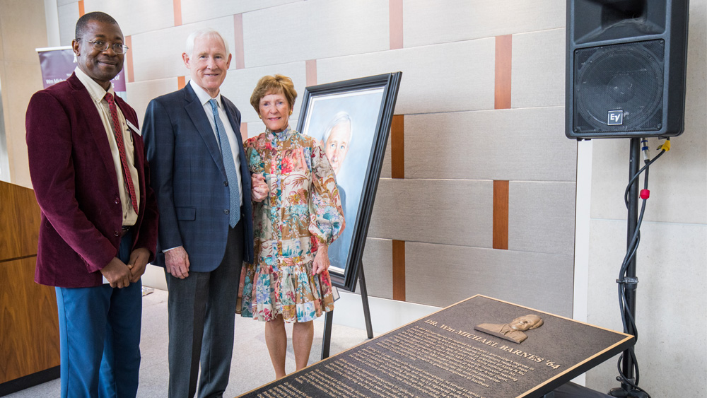 Two men and one woman stand on a platform, smiling at the camera. Next to them is a plaque about the naming of the Wm Michael Barnes '64 Department of Industrial and Systems Engineering.