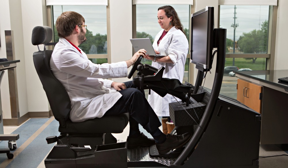 Two researchers in lab coats use a driving simulator in a bright lab, one seated at the wheel and one working on a laptop.