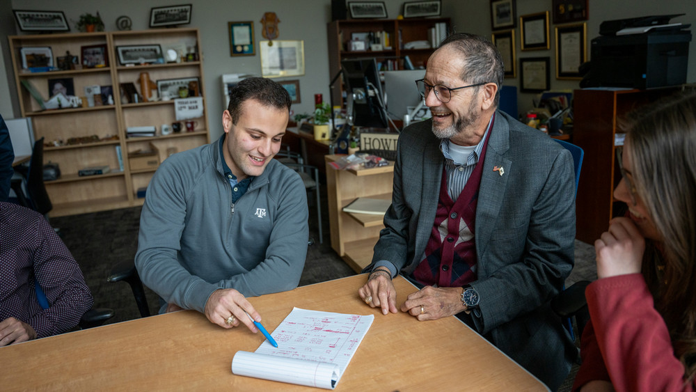 Three people sitting at a table smiling. One is pointing at writing on paper.