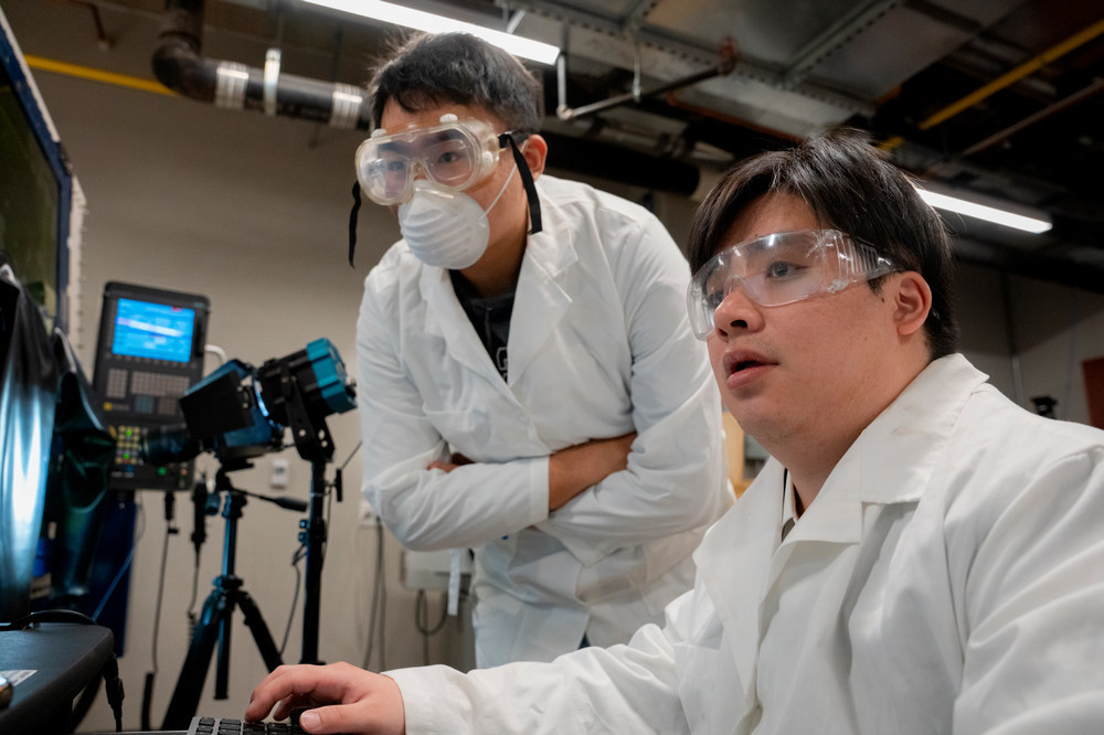 Two students wearing goggles, lab coats and masks starting at a screen in a research lab.