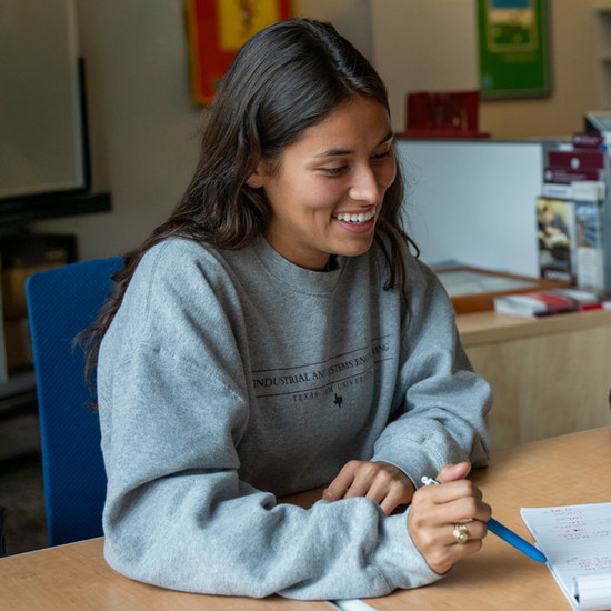 a girl holding a pen, smiling as she points to paper in front of her.