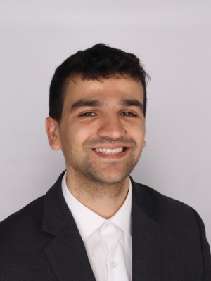 Professional headshot of Rajiv Sambharya, smiling, wearing a dark gray suit jacket over a white collared shirt, against a light gray background.