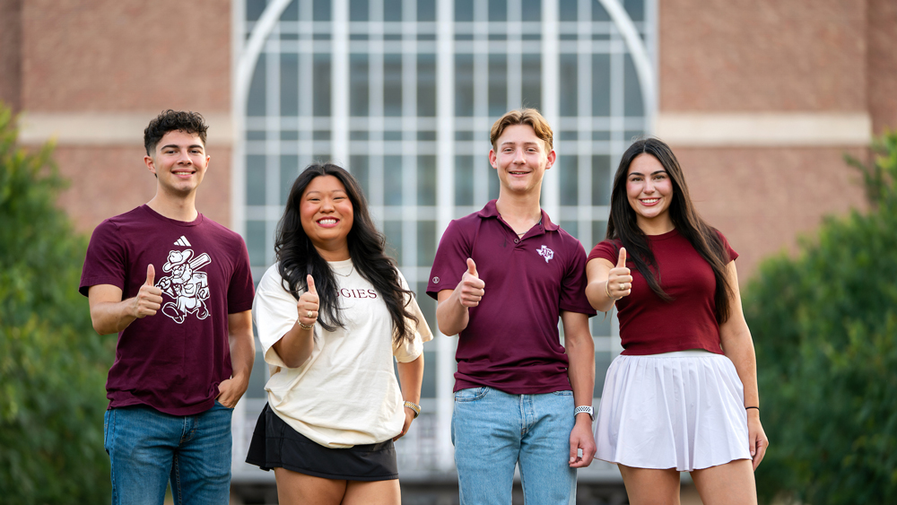  Four diverse, smiling college students stand side by side in front of a large, arched glass window in a brick building, possibly on a university campus. The two students on the ends are wearing maroon shirts (one is a t-shirt with a baseball graphic, the other is a polo), and jeans. The girl second from the left is wearing a tan t-shirt and a black skirt. The girl on the far right is wearing a maroon t-shirt and a white skirt. All four students are giving a thumbs-up sign.