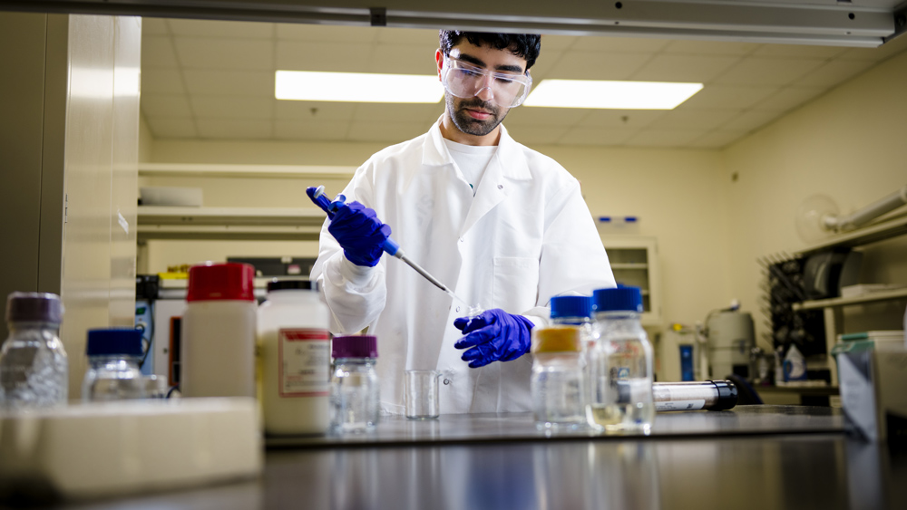 A research student in a white lab coat and safety goggles is working in a lab. He has a dark beard and is wearing blue nitrile gloves. He is using a micropipette to transfer a liquid into a small clear beaker on a stainless steel countertop, surrounded by several bottles and beakers of chemicals and solutions. The background shows lab shelving and ceiling lights.