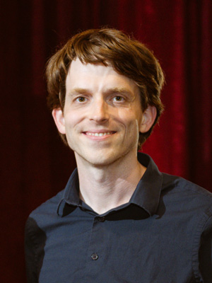 A man smiling with a dark button-up shirt in front of a background. 