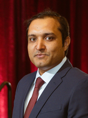Man standing in a suit with a tie in front of a background. 