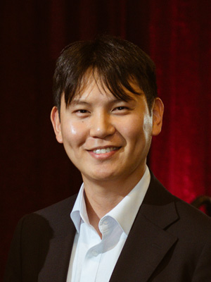 A man smiling with a suit and no tie in front of a background. 