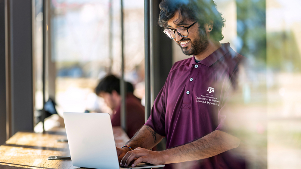 A male college student with a beard and glasses is smiling while working on a silver laptop. He is wearing a maroon polo shirt with the Texas A&amp;M University Department of Materials Science &amp; Engineering logo and is sitting at a table near a large window, possibly in a café or library. Another person is blurred in the background.