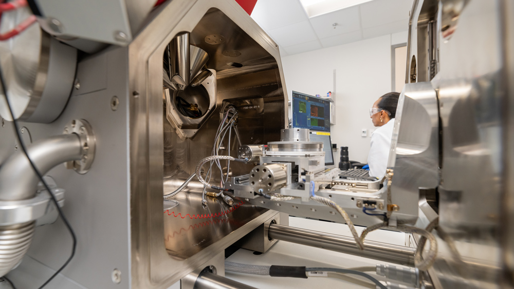 Researcher in a white coat and safety glasses works at a computer in a bright lab, viewed through the open door of a polished steel vacuum chamber filled with cables.