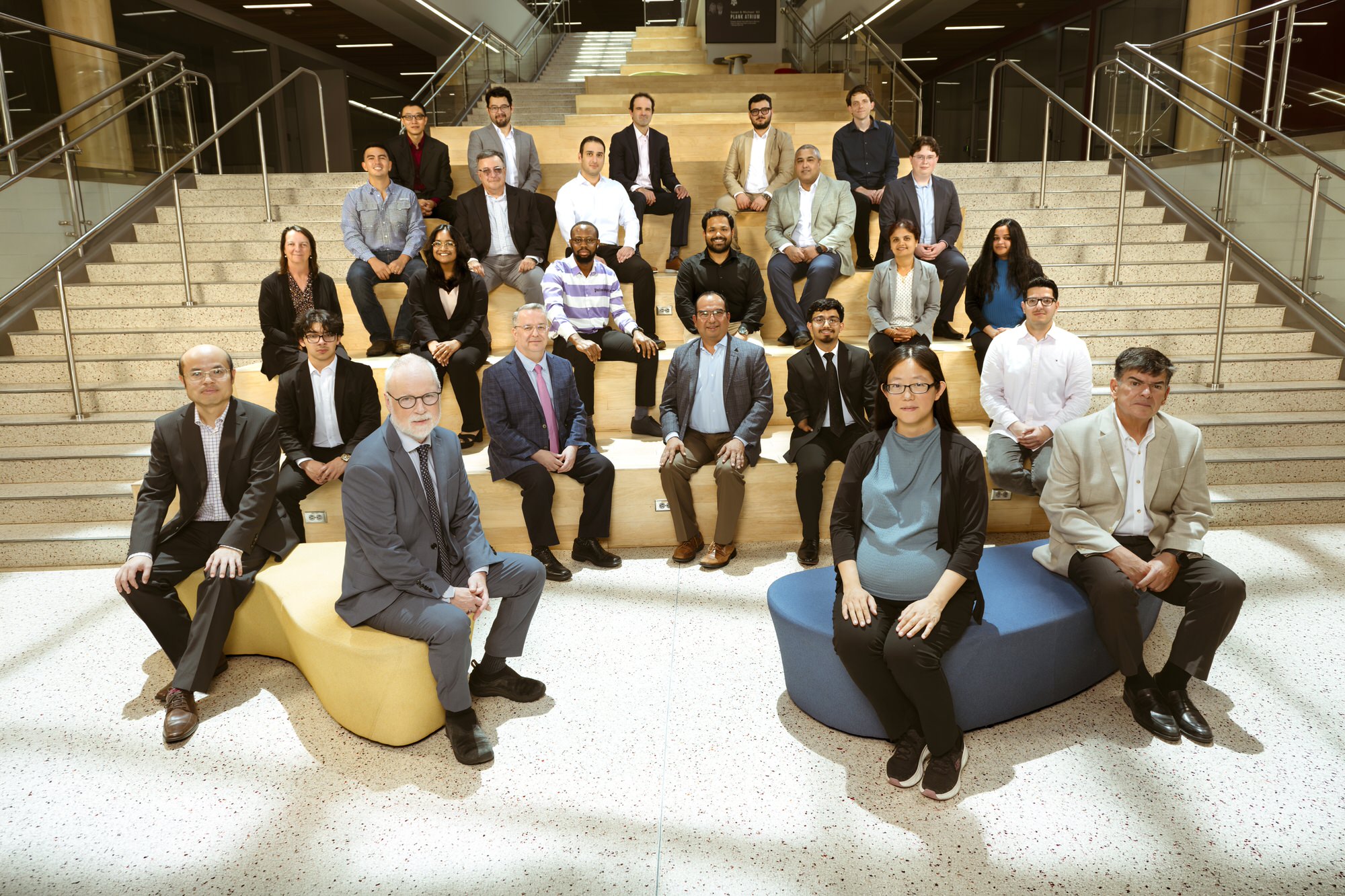 A group of about 25 researchers, faculty, and students pose on the wide wooden staircase and lobby seating of a modern, light-filled campus atrium.
