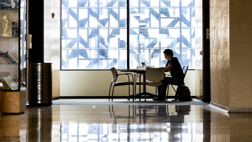 A student in dark clothes studies alone at a small table with a laptop, silhouetted against a large window of geometric patterns, with a backpack on the floor beside them.