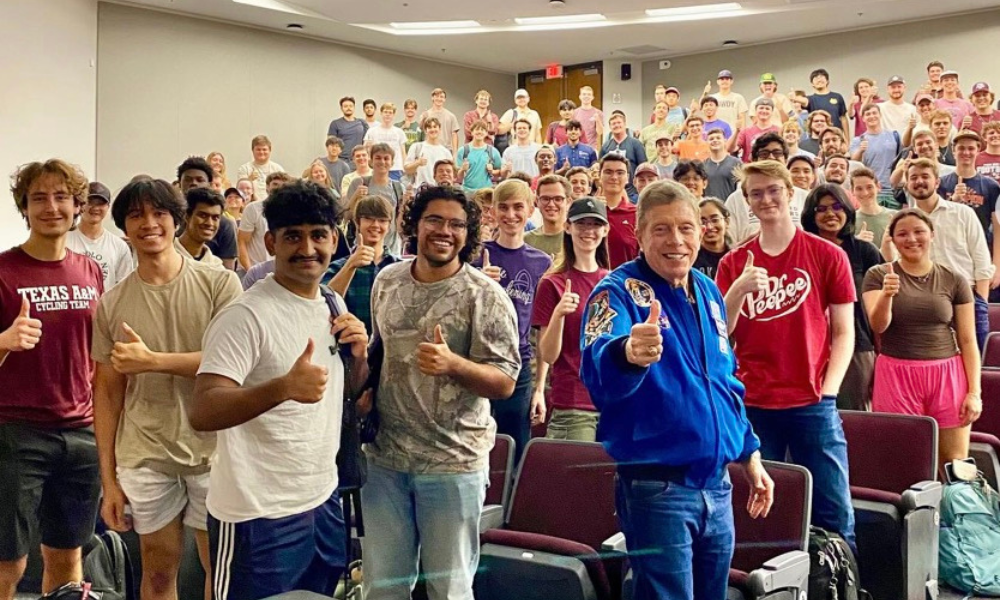 A man in a blue NASA jacket stands in front of a crowd of students in an auditorium. Everyone is holding thumbs up.