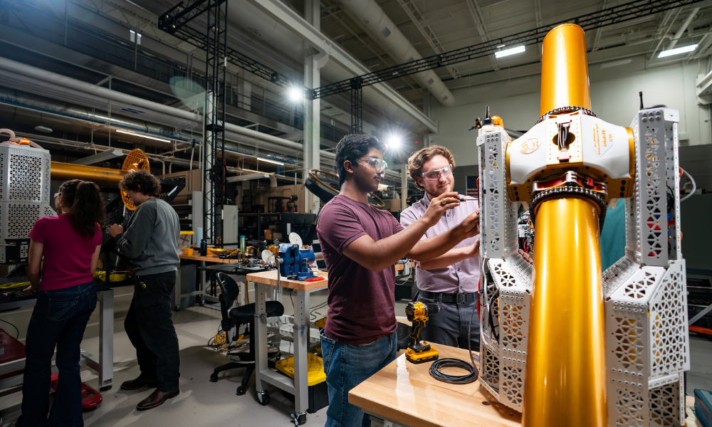 Four students wearing safety goggles work in a large warehouse lab space on large, shiny gold machinery.