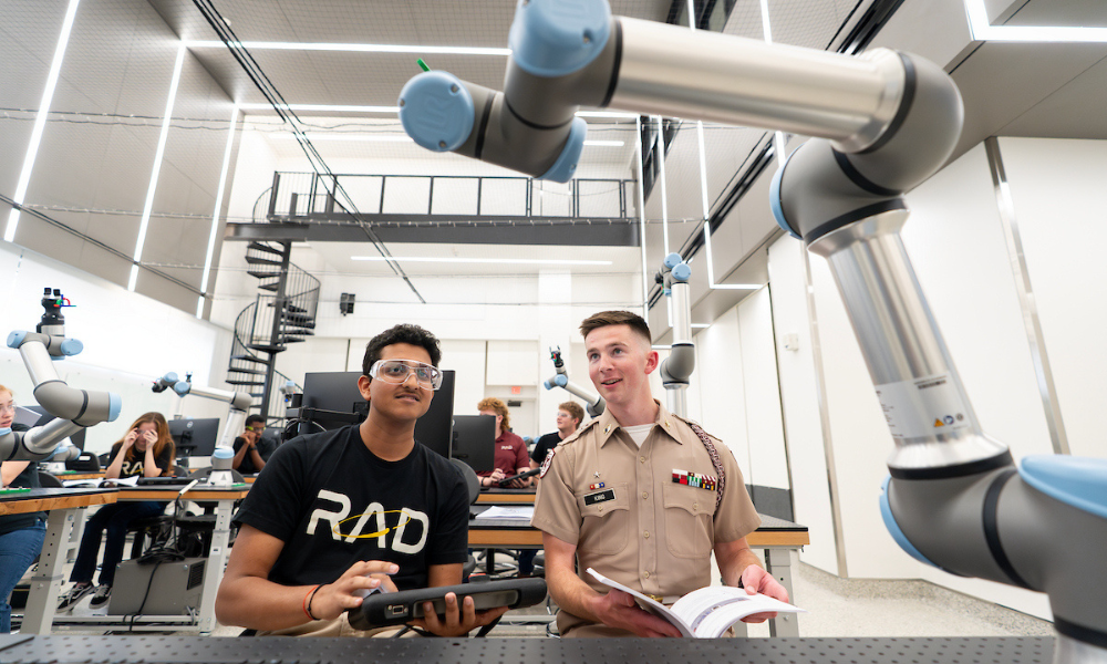 Two students sitting at a desk in a large open lab look up at a robotic arm. One student is wearing safety goggles.