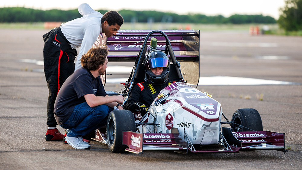 the men are bent over a race car with a driver in the car. the driver is wearing a racing suit and helmet. 