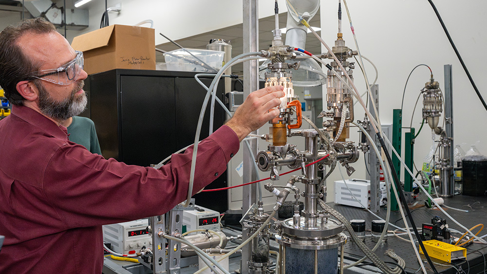 A researcher adjusts a complex laboratory setup with tubes and valves while several students observe in safety glasses