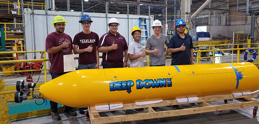 Six students (five male, one female) wearing hardhats standing behind small, yellow submarine giving thumbs up.