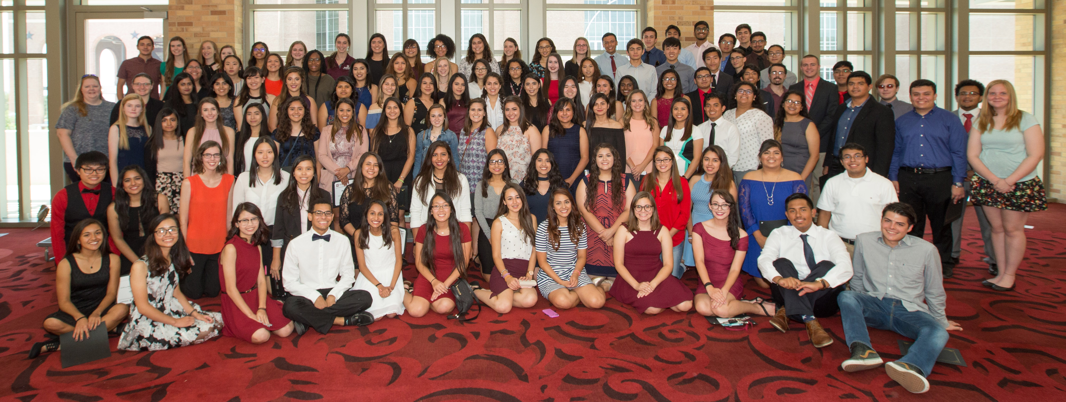 Large group of students posing for photo. all are in business casual clothes. They are in hallway, the carpet is red and behind them is a wall of windows.