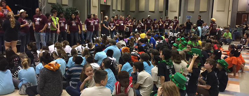 Large group of children sitting in auditorium and the adults are standing in front of them.