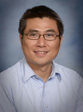 Formal headshot of man with short black hair, wearing glasses with a light blue button up shirt with collar on. The background of the picture is an opaque blue.